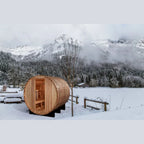 Wooden barrel-shaped sauna in a snowy landscape with mountains and trees.